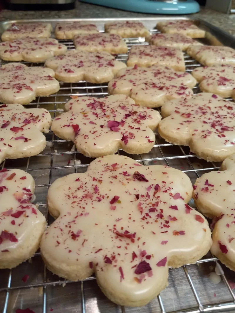 flower-shaped sugar cookies with cream icing and dried rose petals, on a cooling rack